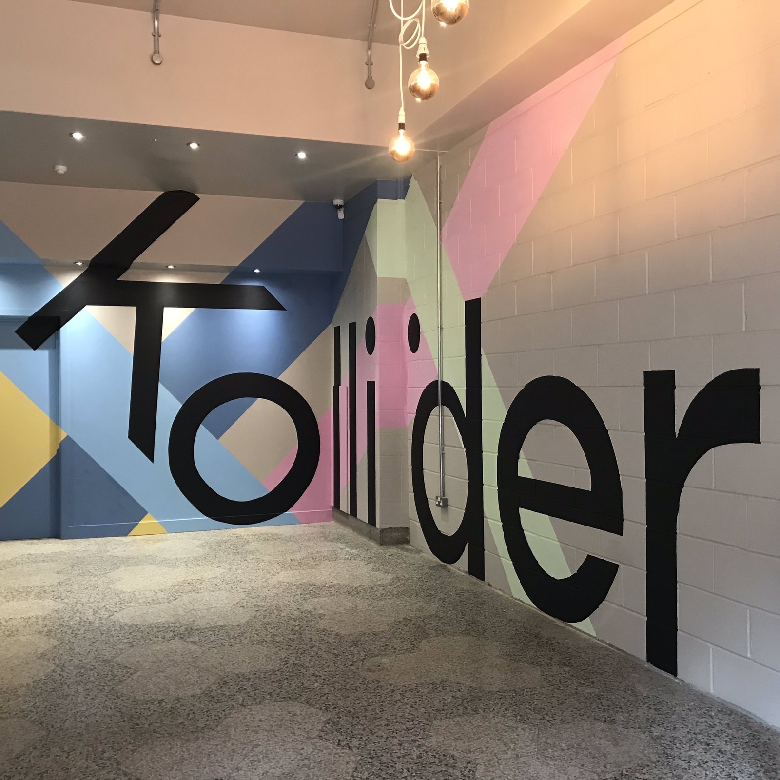 Interior view of a modern hallway with polished terrazzo flooring and colorful geometric wall art. The mural features large black letters spelling “Kollider” across a background of pastel shapes in pink, blue, yellow, and green. Several hanging light bulbs with exposed cords are suspended from the ceiling, adding a contemporary touch.