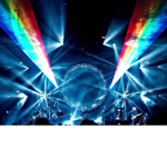 Wide concert stage lit with dramatic blue and white spotlights, featuring a large circular structure at the centre. Rainbow-coloured light beams project diagonally across the venue from both sides. Musicians appear as small silhouettes along the stage, while the audience is visible in shadow at the bottom of the image. The scene creates a striking, immersive live music atmosphere with dynamic lighting effects.