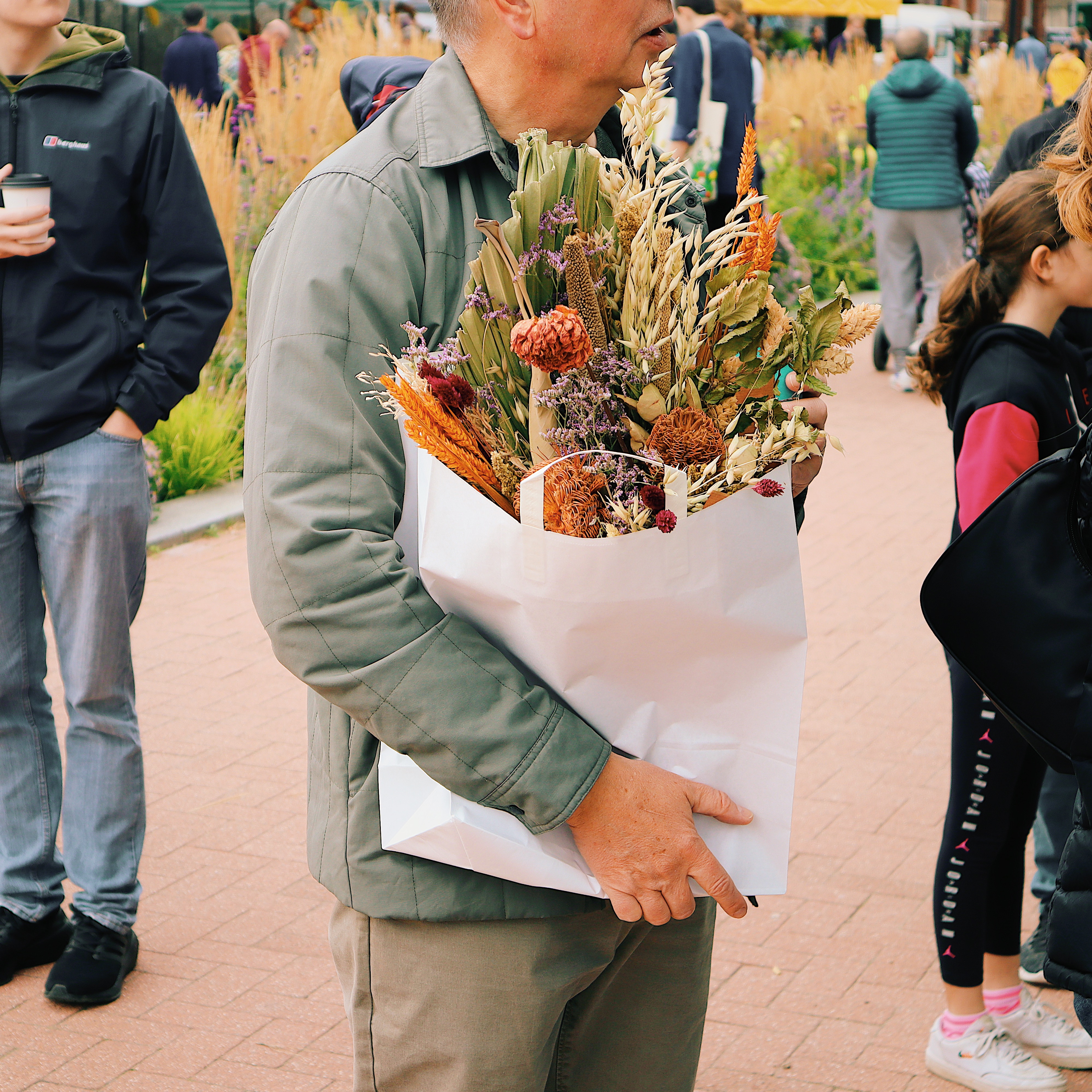 Person holding a large white paper bag filled with dried flowers and foliage, including orange and brown seed pods and stems. The person is wearing a green jacket and standing on a paved walkway with other people and plants visible in the background.
