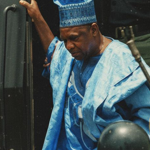 Photograph showing an individual in a blue traditional outfit stepping down from a dark vehicle, surrounded by armed security personnel in military uniforms and helmets. Several people are gathered in the foreground, while one security officer stands beside the vehicle. The scene appears outdoors, with a tense security presence and a large green structure visible to the right.