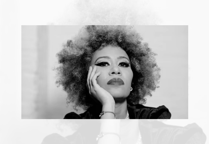 Black-and-white portrait photograph of a person with voluminous curly hair, resting one side of the face on a hand. The image is closely cropped, focusing on the face, hair and upper shoulders. The subject wears earrings and a bracelet, with defined eye makeup visible. A plain, softly lit background keeps attention on the facial features and pose.