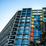 A brutalist high-rise apartment block made of concrete and glass. The windows are interspersed with bright panels in yellows and oranges.