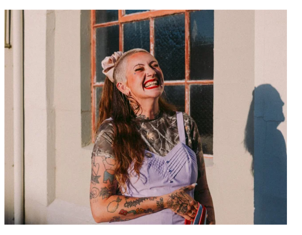 Portrait of a tattooed person standing outdoors in warm sunlight beside a large-paned window. The person wears a patterned top under a light purple pinafore-style dress and holds a striped item in one hand. Long hair is tied back with a large bow. Strong sunlight casts a clear shadow on the wall to the right, while the window and cream-coloured building façade form the background.