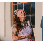 Portrait of a tattooed person standing outdoors in warm sunlight beside a large-paned window. The person wears a patterned top under a light purple pinafore-style dress and holds a striped item in one hand. Long hair is tied back with a large bow. Strong sunlight casts a clear shadow on the wall to the right, while the window and cream-coloured building façade form the background.