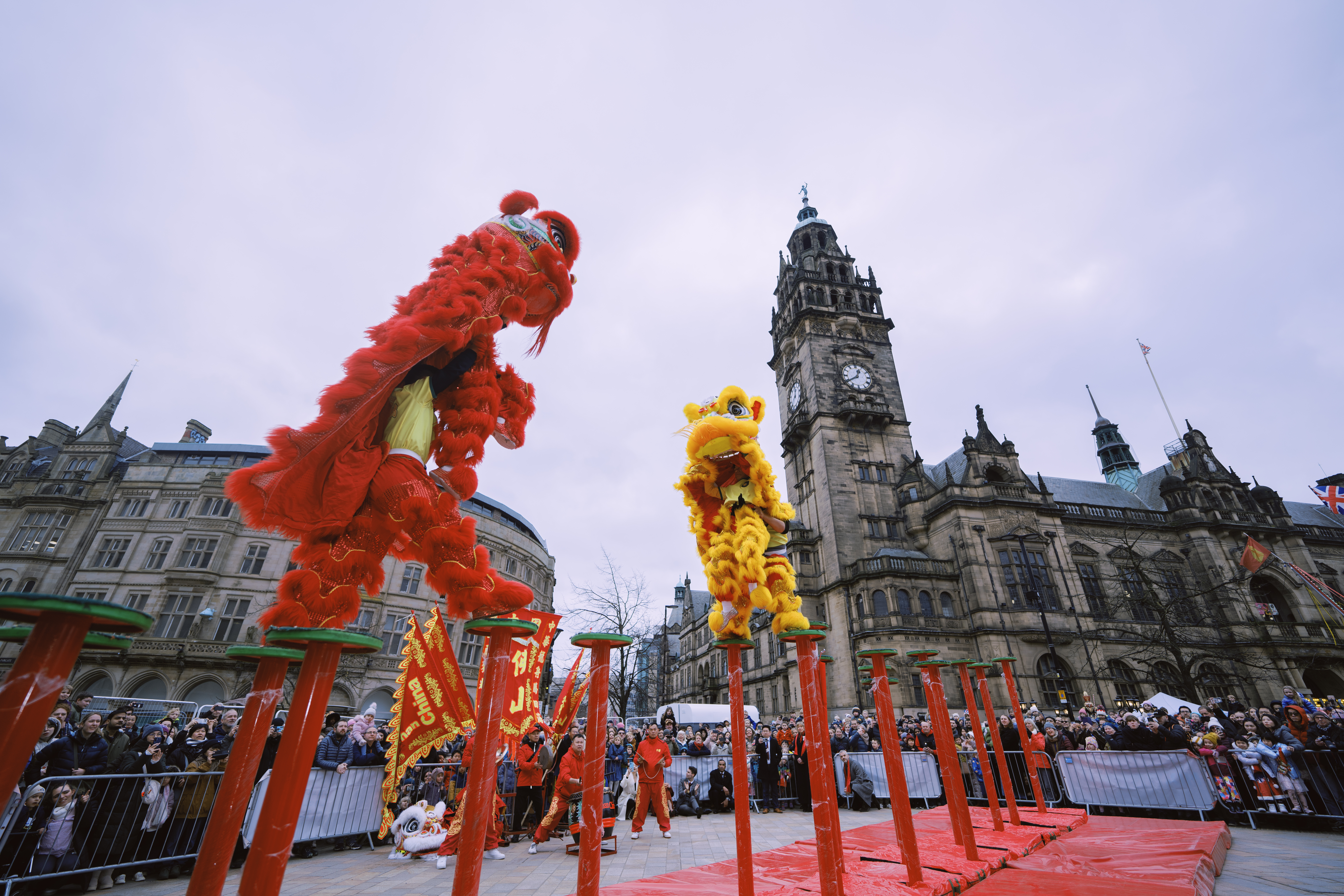 A vibrant lion dance performance in a public square, with two lion costumes—one red and one yellow—balanced on tall poles. A historic clock tower and ornate building dominate the background, and a large crowd surrounds the performance area.