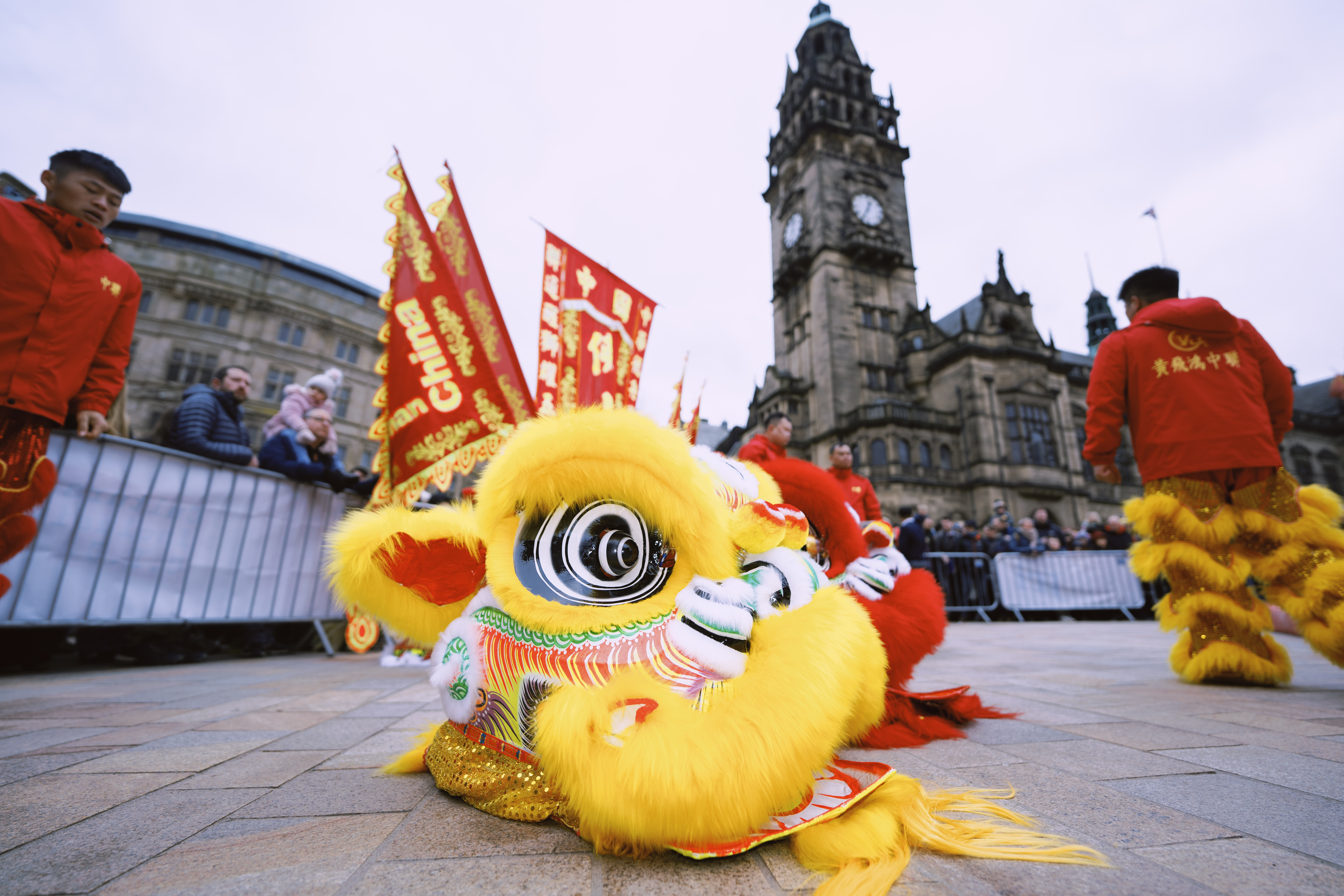 A close-up of a yellow lion dance costume lying on the ground, with intricate detailing and bright colors. Performers in red outfits and banners with Chinese characters are visible in the background, along with a historic building.