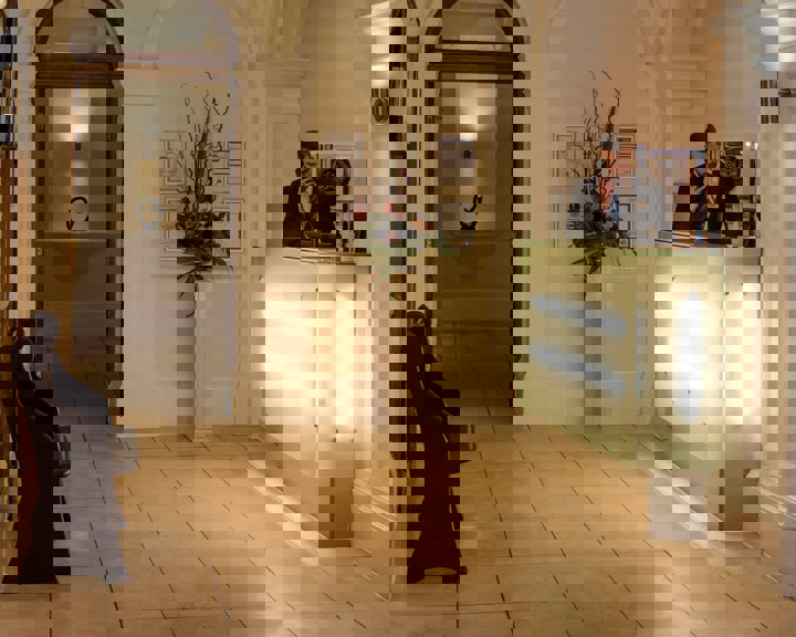 Reception desk in the Leopold Hotel - Reception area of a hotel with a modern glass-front desk illuminated by soft lights. Behind the desk are framed certificates and a tall floral arrangement. The space features cream-colored walls, arched doorways, and tiled flooring. A wooden bench is positioned along the left wall, and an open doorway in the background reveals a decorative sculpture on a shelf.