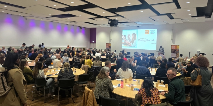 A large conference room filled with round tables where attendees are seated and engaged in discussion. The room has a modern design with a wooden floor, white walls, and a ceiling featuring black acoustic panels. Purple accent lighting runs along the left wall. At the front of the room, a large screen displays a presentation titled “Celebrating Culture & Creativity” with the James Reed logo. A speaker stands near the screen, and event banners are positioned on both sides of the stage area.