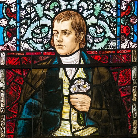 Stained glass artwork depicting a figure in period clothing holding a small bouquet of white daisies. The figure wears a dark outer garment over a light waistcoat and shirt. The background features intricate patterns in shades of green, blue, and red, framed by ornate designs, creating a rich and detailed composition.
