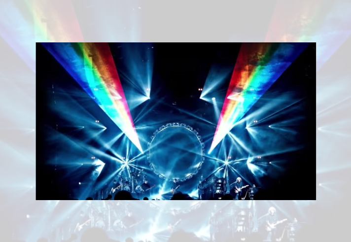Wide concert stage lit with dramatic blue and white spotlights, featuring a large circular structure at the centre. Rainbow-coloured light beams project diagonally across the venue from both sides. Musicians appear as small silhouettes along the stage, while the audience is visible in shadow at the bottom of the image. The scene creates a striking, immersive live music atmosphere with dynamic lighting effects.