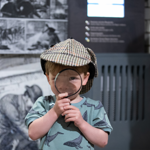 Child wearing a Sherlock Holmes-style deerstalker hat holds a magnifying glass in front of one eye while standing inside a museum exhibit. In the background are blurred black-and-white Victorian crime investigation illustrations and display panels, reinforcing a detective and mystery theme.