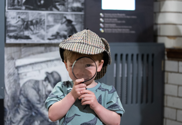 Child wearing a Sherlock Holmes-style deerstalker hat holds a magnifying glass in front of one eye while standing inside a museum exhibit. In the background are blurred black-and-white Victorian crime investigation illustrations and display panels, reinforcing a detective and mystery theme.