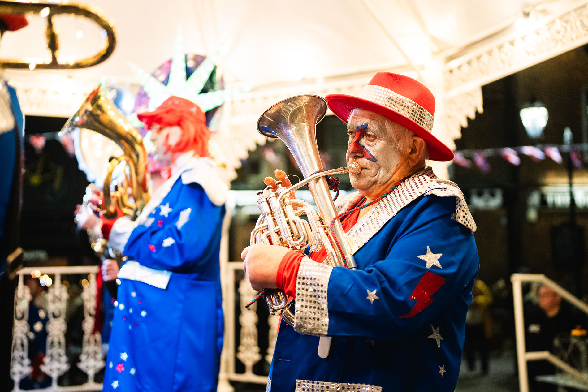 Two performers dressed in bright blue costumes decorated with silver stars and red lightning bolts play brass instruments under a white ornate gazebo. One performer wears a red hat and holds a shiny silver euphonium, while another stands slightly behind, also playing a brass instrument. The scene is well-lit, suggesting a festive outdoor event at night, with decorative lights and bunting visible in the background.