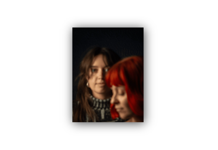 Portrait of two women against a dark background. One woman with bright red hair is in the foreground, shown in soft focus and looking downward, while a second woman with brown hair stands behind her, partially in focus and looking towards the camera. The image uses shallow depth of field and warm lighting to create an artistic, intimate composition.