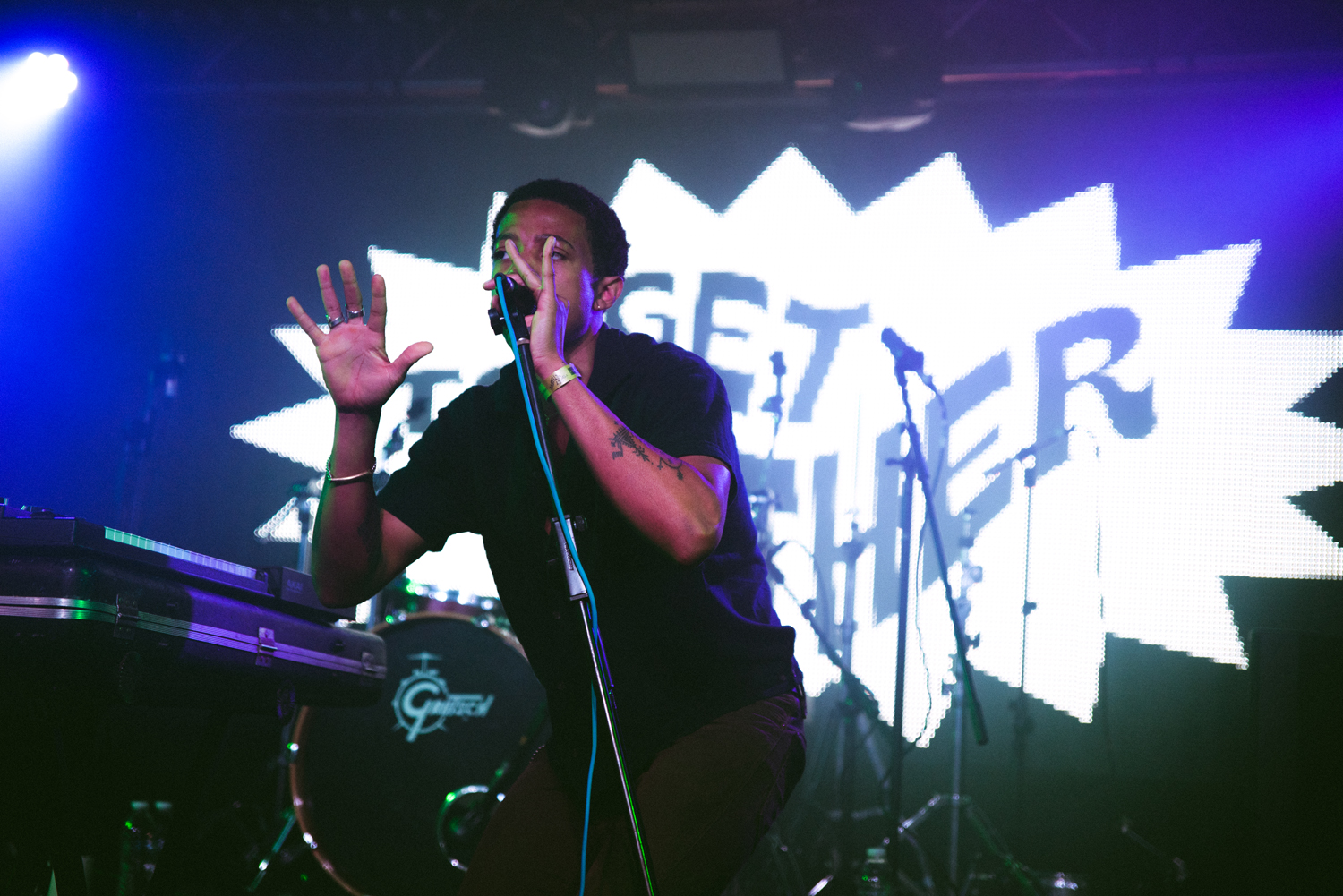 A performer on stage holding a microphone and gesturing with one hand, standing behind a DJ-style setup with equipment. The background features a large illuminated graphic with bold text reading “GET TOGETHER” in a comic-style burst design. The stage is lit with vibrant blue and purple lighting, and drum kits and microphone stands are visible behind the performer, creating a lively concert atmosphere.