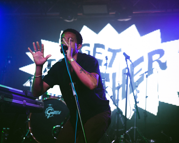 A performer on stage holding a microphone and gesturing with one hand, standing behind a DJ-style setup with equipment. The background features a large illuminated graphic with bold text reading “GET TOGETHER” in a comic-style burst design. The stage is lit with vibrant blue and purple lighting, and drum kits and microphone stands are visible behind the performer, creating a lively concert atmosphere.