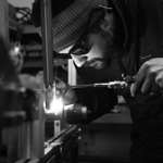 A black and white photo of a man welding a bike frame in a workshop.
