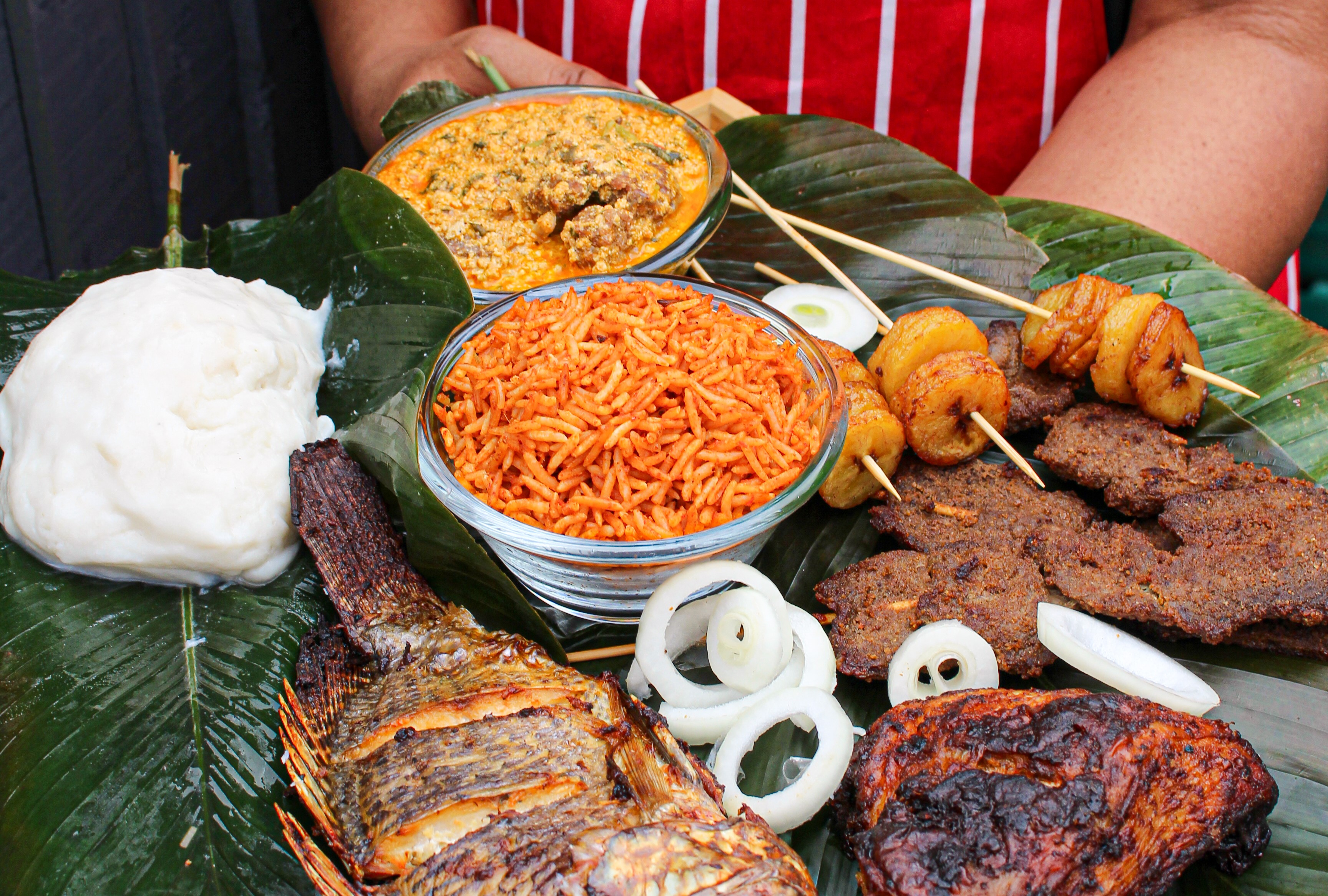 Assorted traditional African dishes arranged on large green leaves. The spread includes grilled fish, grilled chicken, skewered fried plantains, skewered spiced meat, a bowl of jollof rice, a bowl of groundnut stew, sliced onions, and a portion of white starchy paste (likely fufu).
