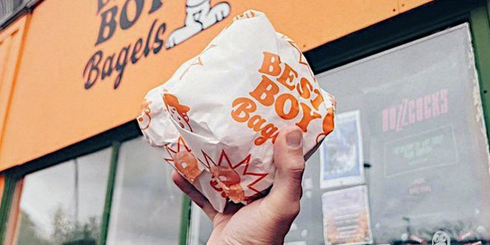A hand holds a wrapped bagel from Best Boy Bagels, whose sign can be seen in the background.