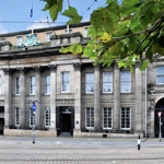 Exterior view of a grand historic stone building with a neoclassical façade, featuring tall fluted columns, large sash windows and decorative architectural details. Tram tracks and overhead wires run along the street in the foreground, while green tree branches and leaves frame the top right of the image against a partly cloudy sky.