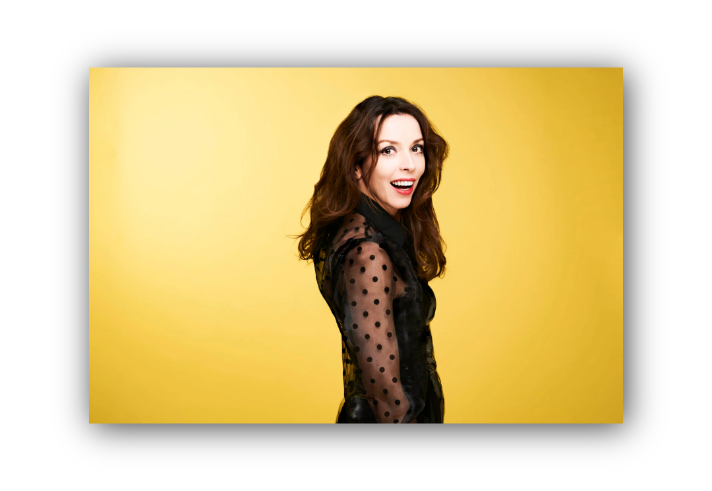 Person standing against a bright yellow background, wearing a sheer black top with polka-dot sleeves and solid black panels. The individual has long, wavy brown hair and is posed in profile, facing slightly away from the camera.