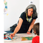 A workshop facilitator leans over a table demonstrating printmaking techniques to a child seated opposite. Art materials including rollers, ink trays and printed papers are spread across a covered work surface. The participant wears a yellow lanyard and reaches toward a roller coated with blue ink. The bright room has a plain white wall, with a colour wheel poster partly visible in the background.