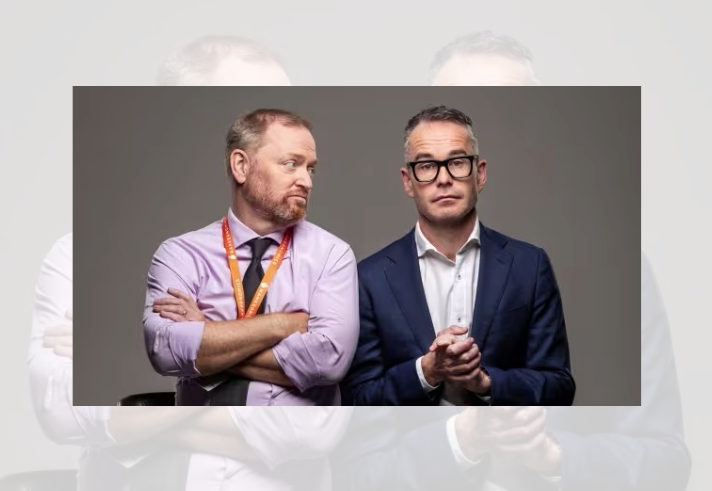 Studio portrait of two people seated side by side against a plain grey background. One wears a light purple shirt with an orange lanyard and sits with arms folded, looking towards the other person. The second wears a navy suit jacket, white shirt and dark-framed glasses, with hands clasped in front. The image has a clean, professional style with neutral lighting and no visible text.