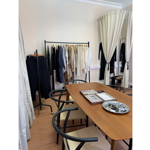 Interior of a minimalist clothing showroom with pale walls, wooden flooring and cream curtains. A clothing rail displays neatly arranged garments in black, navy, beige and white tones, while a separate stand holds a pair of dark trousers. In the foreground, a wooden table surrounded by black-framed chairs displays jewellery trays and a metal dish with accessories. A standing mirror and additional clothing are visible in the background, creating a calm, curated retail setting.