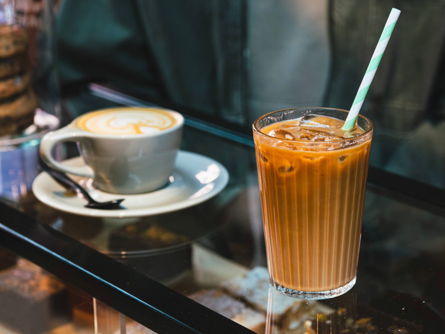 A close-up of two drinks on a glass counter in a coffee shop: a grey ceramic cup of latte with foam art on a saucer and spoon, and a tall glass of iced coffee with a green-and-white striped paper straw. Behind the drinks, part of a person’s jacket is visible, and jars of biscuits can be seen in the background.