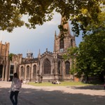 Sheffield Cathedral viewed from a shaded square, with large trees framing the scene. The historic stone building features a tall spire, ornate windows, and Gothic architecture. People are walking and sitting on benches in the sunlit open space in front of the cathedral.