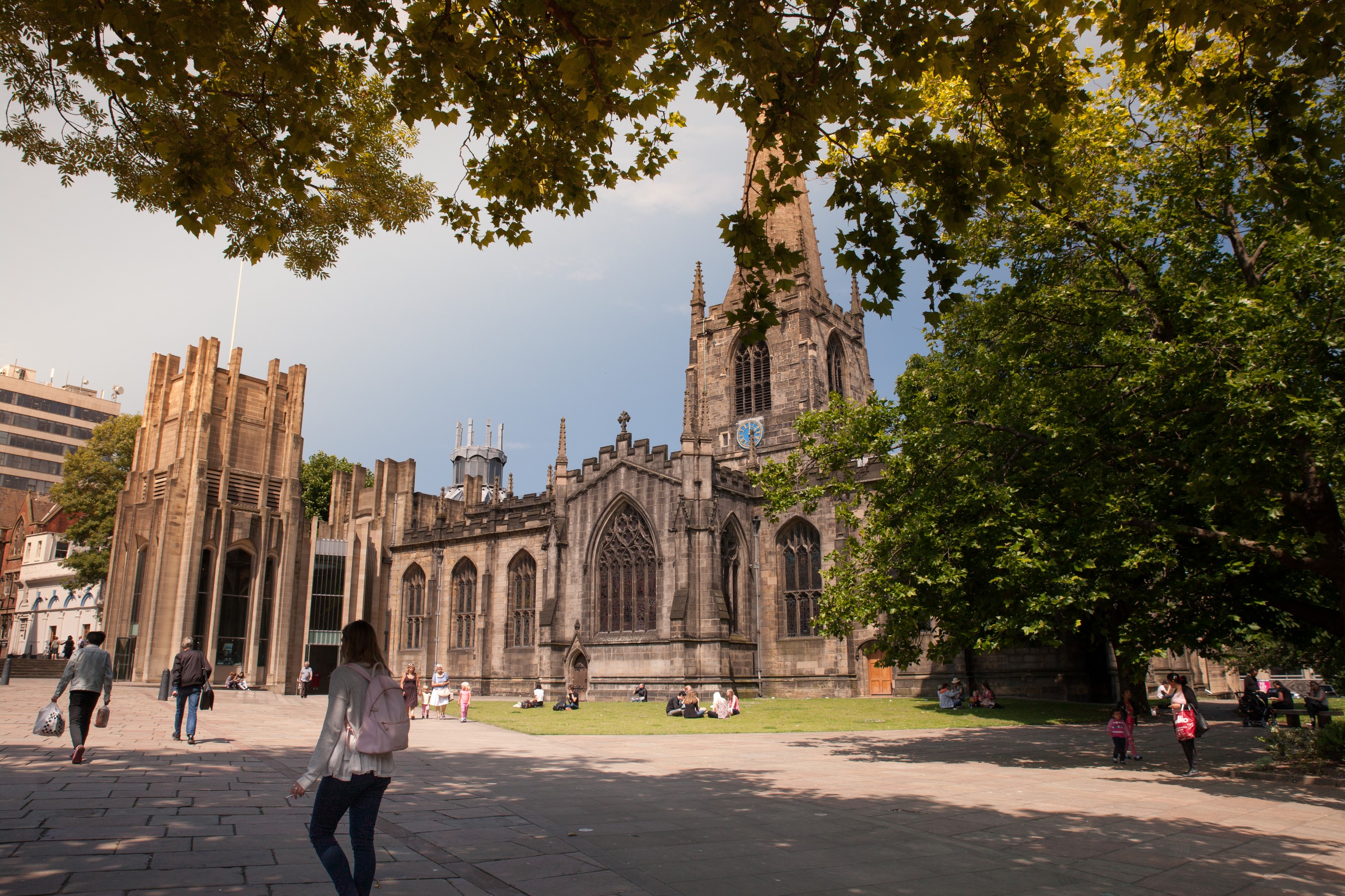 A historic stone cathedral with Gothic architectural features, including pointed arches, tall windows, and a central tower with a clock. The building is surrounded by a paved square with people walking and sitting on the grass. Large trees frame the scene, and sunlight filters through the leaves, casting shadows on the ground. Modern buildings are partially visible in the background.