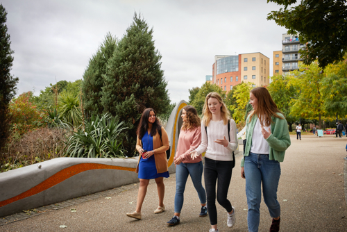 Four people walking along a paved pathway in an outdoor park setting, surrounded by greenery and trees. Modern apartment buildings are visible in the background, and the group appears to be engaged in conversation. The scene is bright and colourful, with a mix of casual clothing and autumn foliage.