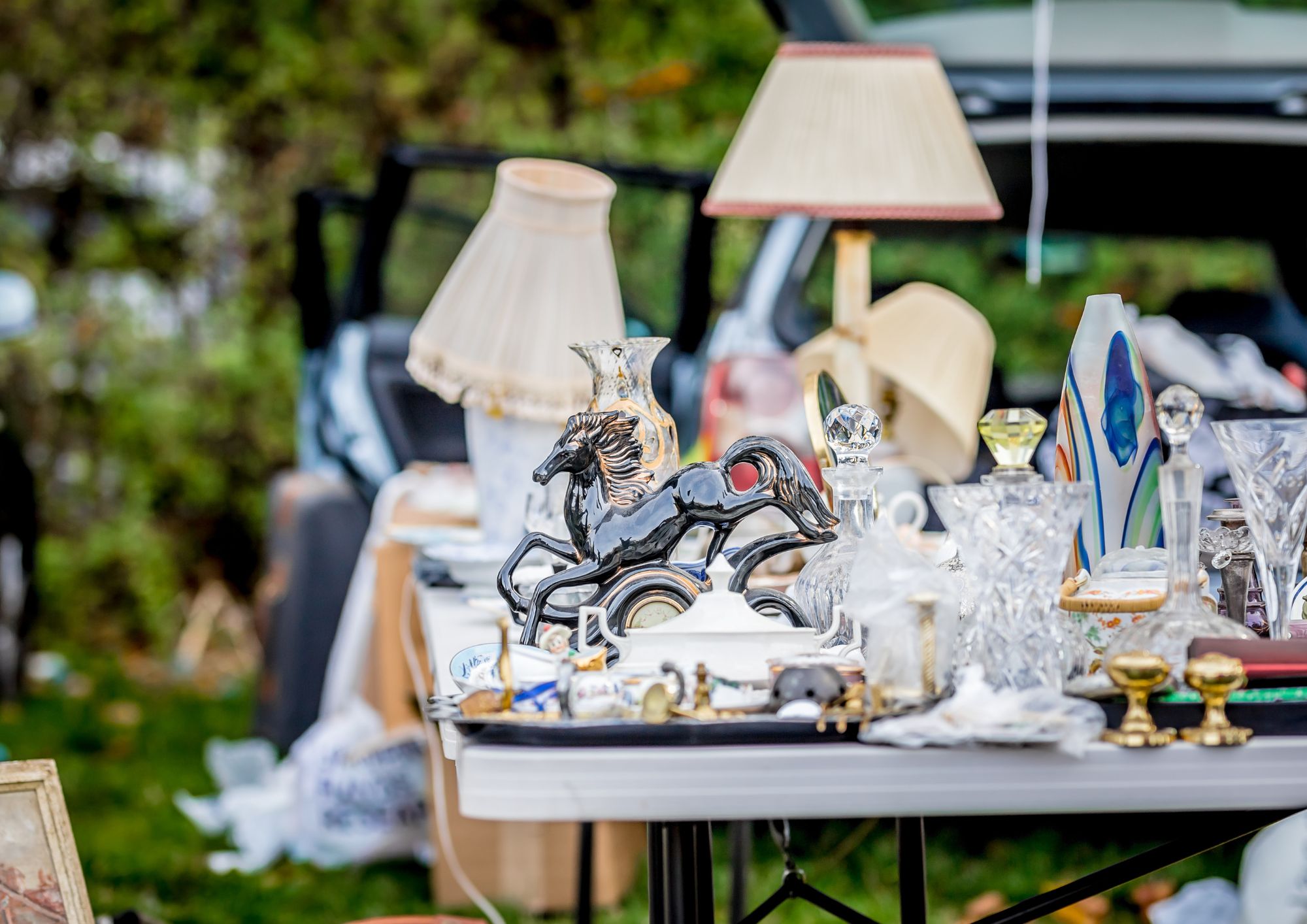 A table at an outdoor car boot sale covered with assorted second-hand items, including a black horse figurine, glassware, brass ornaments, lamps with fabric shades, and decorative vases. More items and an open car boot are visible in the background, with greenery behind them.