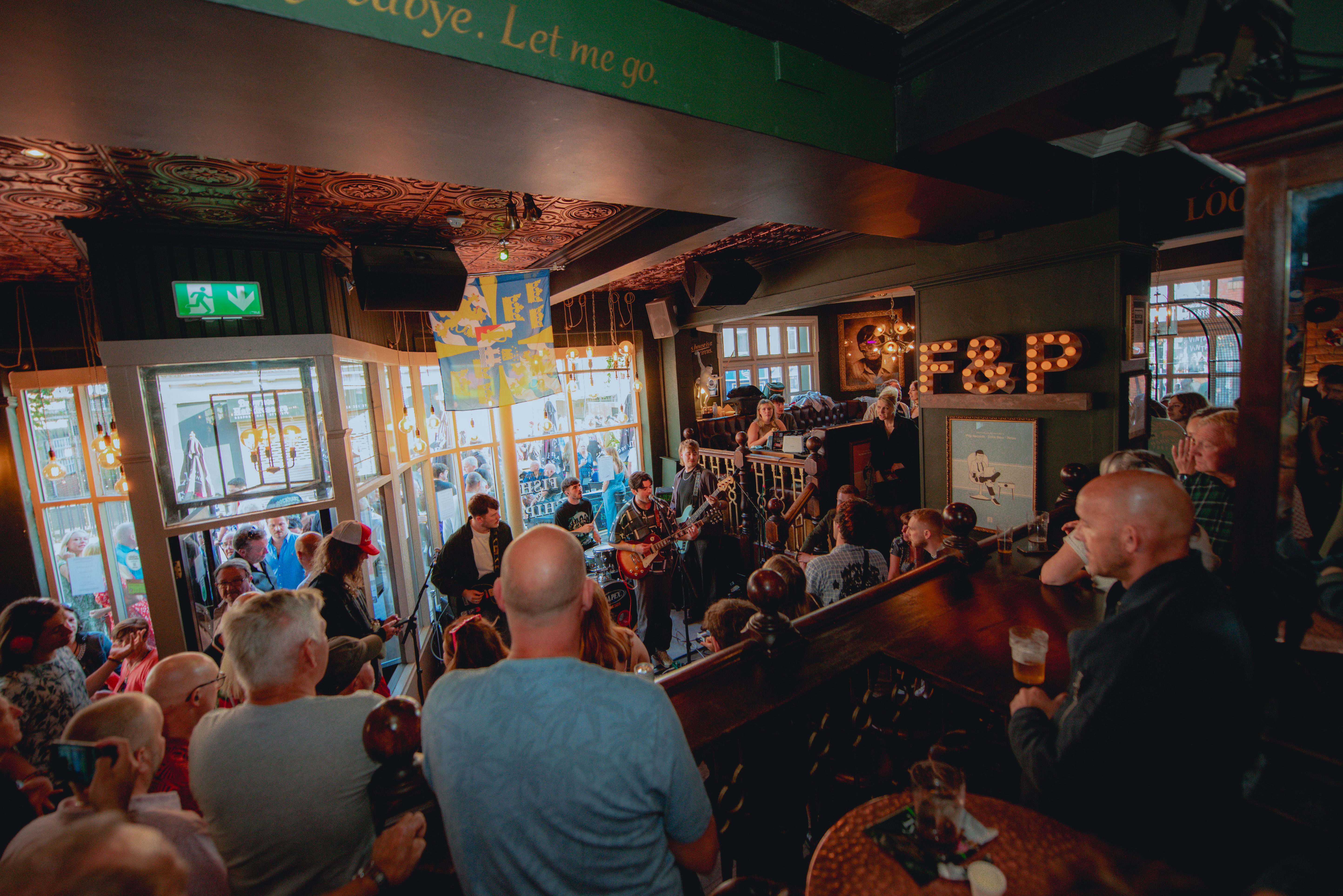 A crowded live music performance inside the Frog & Parrot pub, with warm lighting, patterned ceilings and a band playing near the front windows while people gather closely to watch.