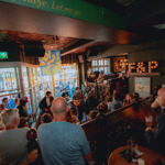 A crowded live music performance inside the Frog & Parrot pub, with warm lighting, patterned ceilings and a band playing near the front windows while people gather closely to watch.