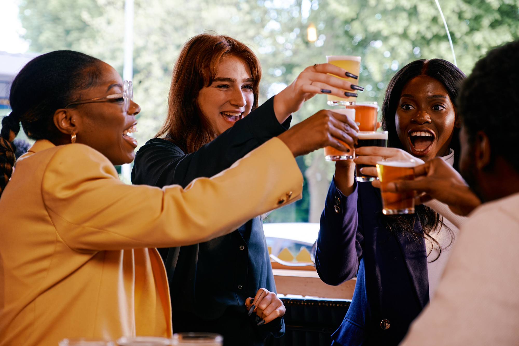 A group of people raising glasses of beer and juice in a toast inside a bright, casual setting with large windows and greenery visible outside. The individuals are wearing business-casual attire, and the scene conveys a celebratory or social gathering.