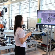 A woman is standing in front of a monitor and holding a tablet. In the background is an industrial robot arm.