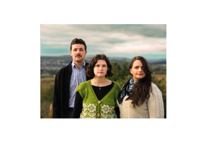 Three people stand outdoors on a hillside with a blurred landscape and cloudy sky in the background. The person in the centre wears a green knitted cardigan with a white floral pattern over a dark top. The person on the left wears a light shirt and dark jacket, while the person on the right wears a cream jumper and patterned scarf. All three face the camera in a posed group portrait.