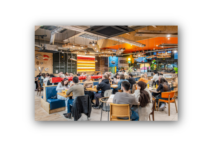 Busy modern restaurant interior with exposed industrial-style ceiling ducts, suspended wooden light fixtures and colourful décor. Numerous diners sit at tables and booth seating across a large open-plan space. Warm lighting, a wall of illuminated bottles and large windows create a lively atmosphere, while motion blur captures the movement and energy of a bustling evening service.