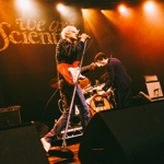 Live performance by rock band We Are Scientists on a theatre stage. A guitarist and vocalist with a red electric guitar performs at a microphone under orange stage lights, while another musician kneels beside equipment near the drum kit. Large black stage monitors fill the foreground, amplifiers sit at the sides, and the band's name, “We Are Scientists”, is displayed in large lettering behind the performers.