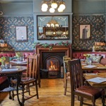  A warm, vintage‑style pub dining area with patterned wallpaper, red leather booth seating, wooden tables, and a lit fireplace beneath a decorative mirror.