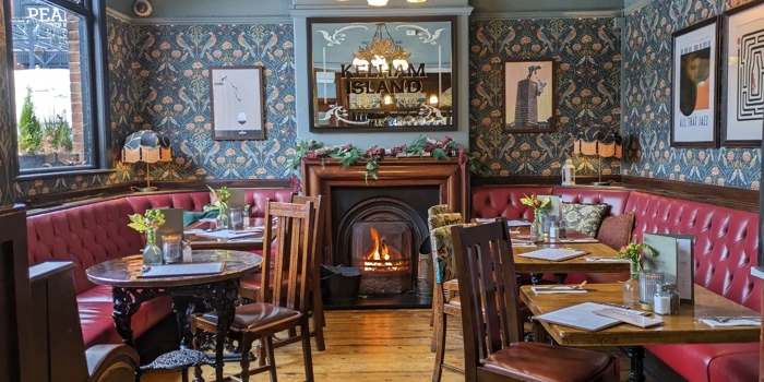  A warm, vintage‑style pub dining area with patterned wallpaper, red leather booth seating, wooden tables, and a lit fireplace beneath a decorative mirror.