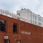 Exterior of the red brick SITE Gallery building with the gallery name displayed on the wall and an 'everything is different today' illuminated sign and cloudy sky above.