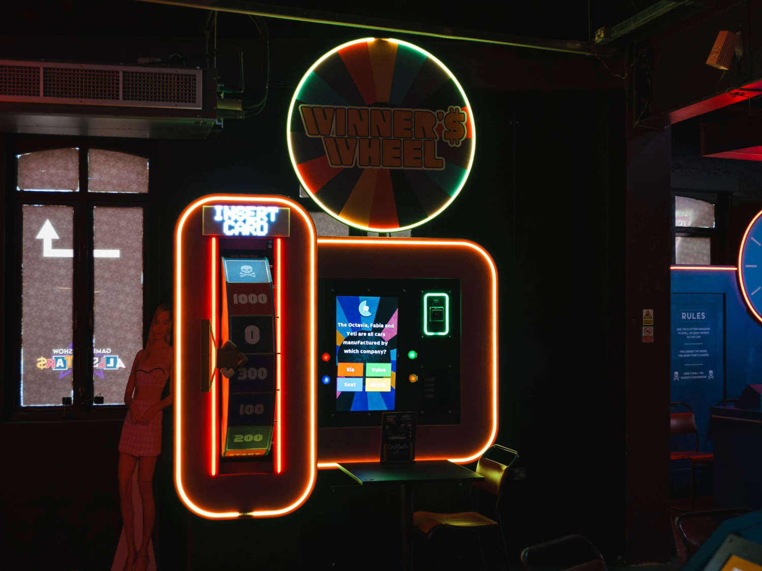 Neon-lit arcade machine called “Winner’s Wheel” with a colourful circular sign above and bright orange framing. The machine features a vertical panel with “Insert Card” text and score markers from 200 to 1000, alongside a digital screen displaying game instructions. A small table and chair sit in front of the machine, and a cardboard cut-out figure is partially visible to the left. The setting is dark with vibrant lighting, creating a lively atmosphere.