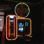 Neon-lit arcade machine called “Winner’s Wheel” with a colourful circular sign above and bright orange framing. The machine features a vertical panel with “Insert Card” text and score markers from 200 to 1000, alongside a digital screen displaying game instructions. A small table and chair sit in front of the machine, and a cardboard cut-out figure is partially visible to the left. The setting is dark with vibrant lighting, creating a lively atmosphere.