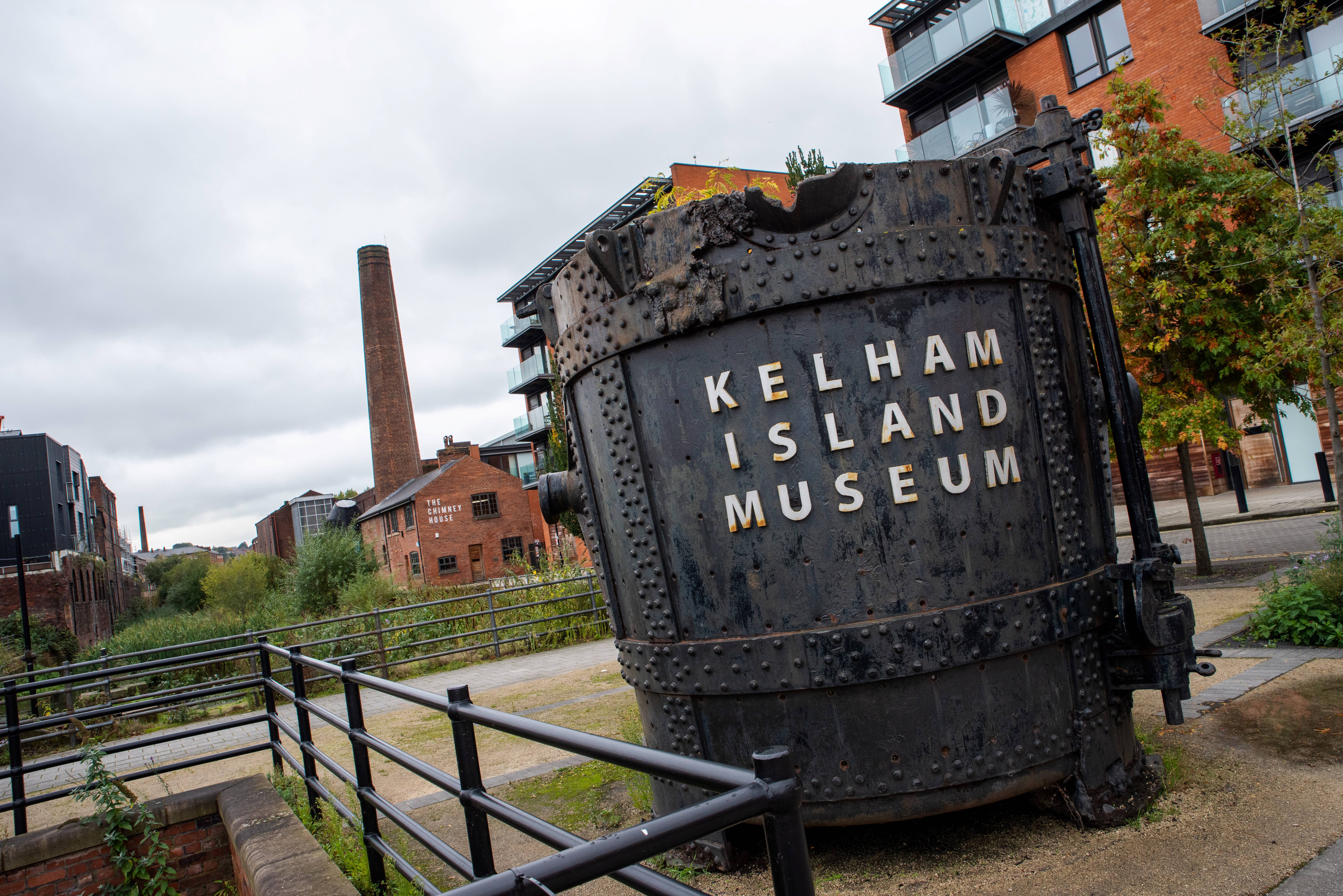 Large industrial steel vessel with rivets and the words “Kelham Island Museum” in bold letters, displayed outdoors on a paved path. Surrounding area includes railings, trees, and modern brick buildings, with a tall chimney visible in the background under a cloudy sky.