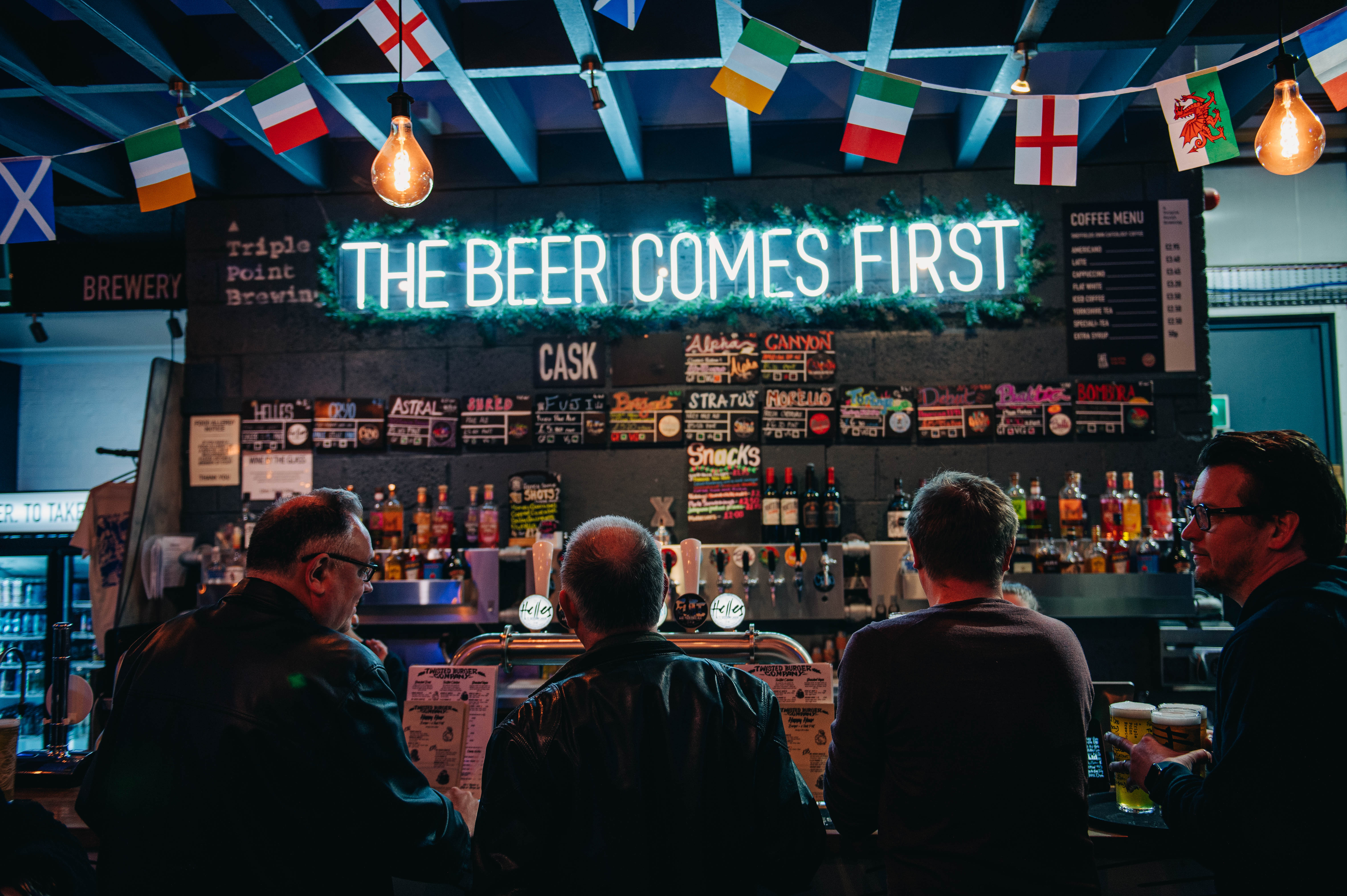 Customers being served at a busy bar with an illuminated sign overhead that reads "the beer comes first"