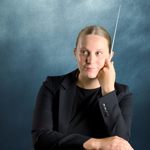A studio portrait of a person in a dark suit holding a conductor's baton.