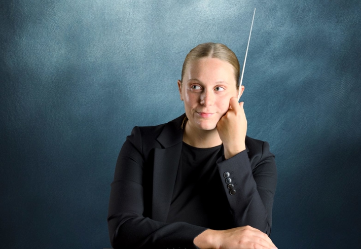 A studio portrait of a person in a dark suit holding a conductor's baton.