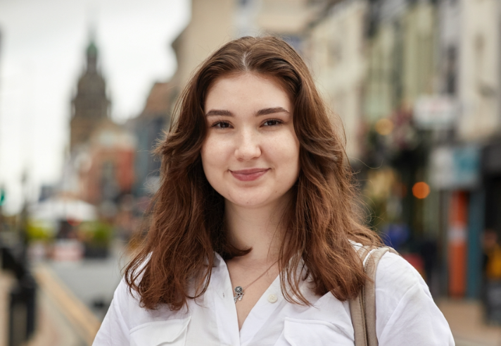 A person wearing a white button-up shirt with a necklace and a shoulder bag, standing on a city street. The background shows a row of buildings with shops and a distant clock tower, along with blurred street details and warm lighting.